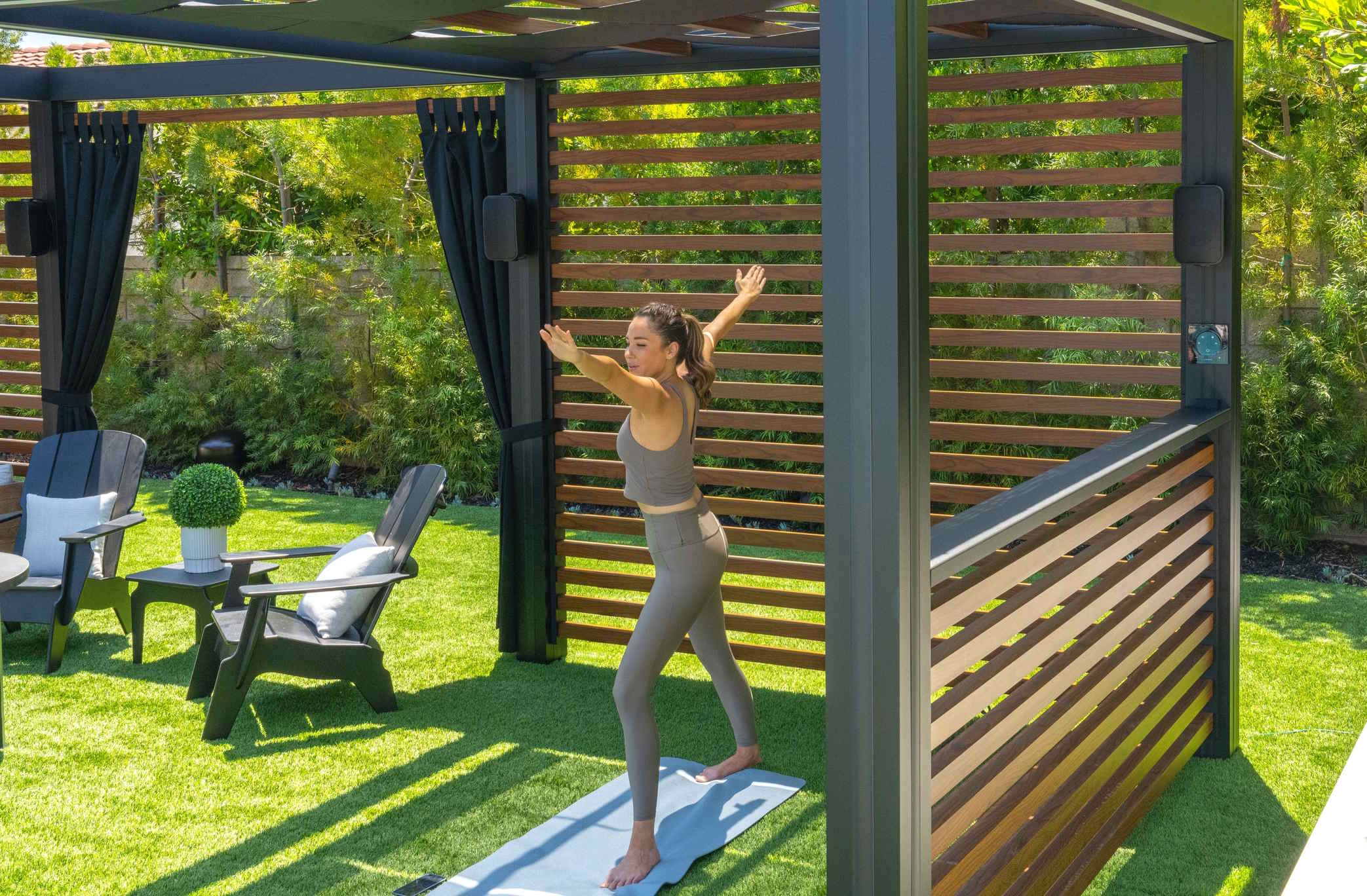 Outdoor gazebo with wooden slats where a person practices yoga on a mat in a landscaped backyard.