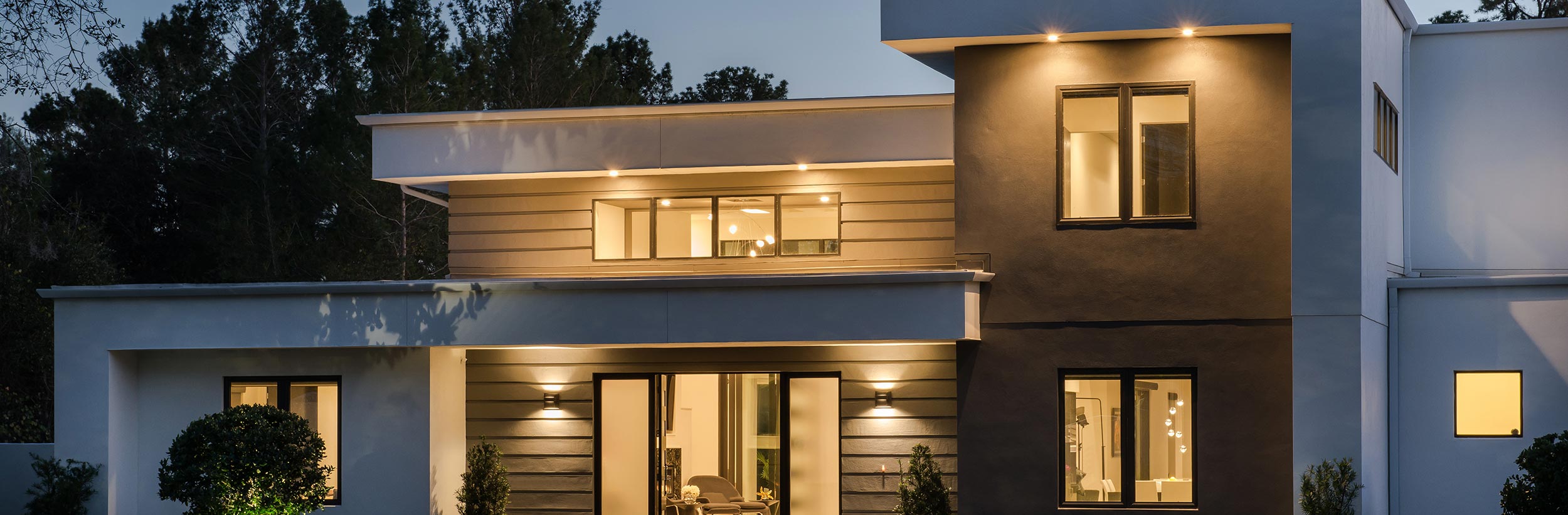Modern two-story home at dusk with warm interior lighting, flat rooflines, and a sleek facade combining light and dark finishes.